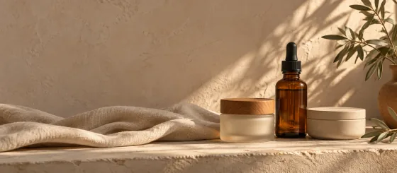 Three unbranded skincare vessels — a frosted jar with a wooden lid, an amber dropper bottle and a pale ceramic tub — grouped on a plaster ledge with cream linen and an olive sprig.